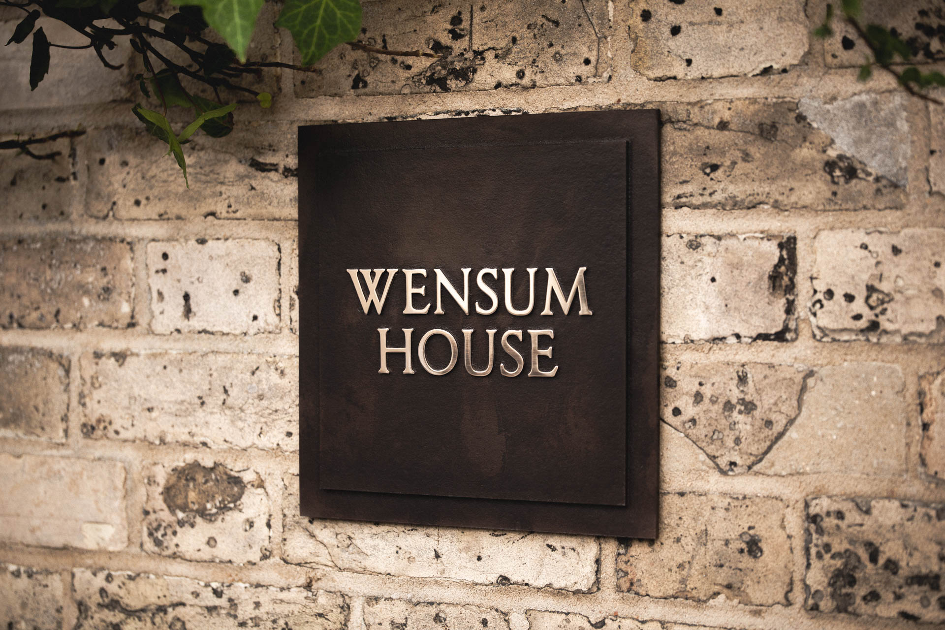 Cast bronze house sign reading “Wensum House” with raised polished bronze lettering, dark patinated background, and two-tiered design, mounted on a light weathered brick wall with green ivy above.