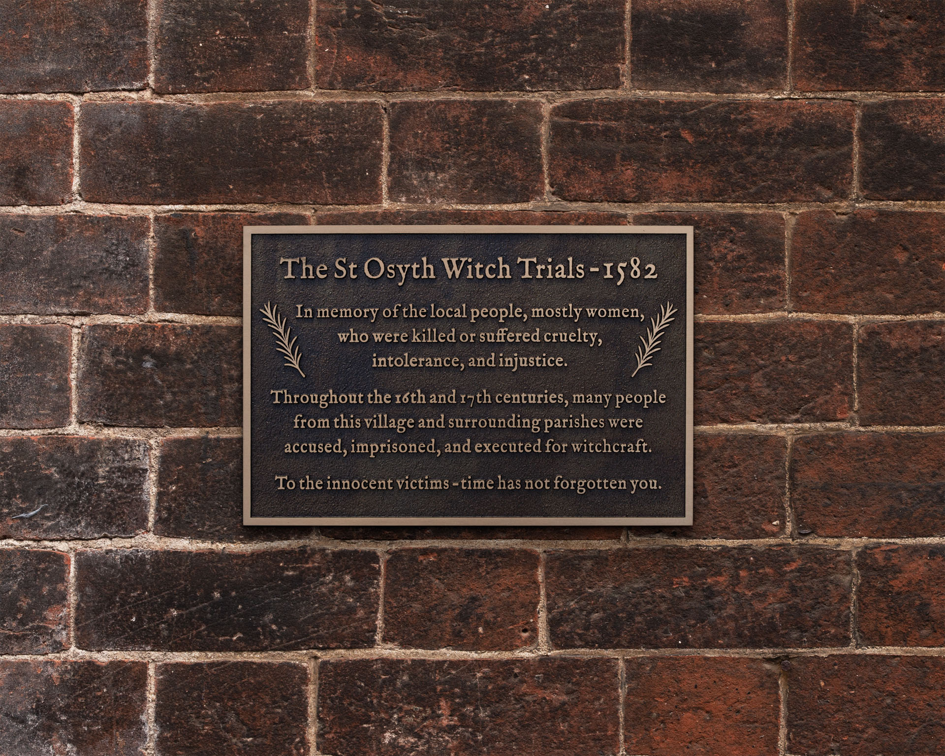 Cast bronze remembrance plaque commemorating the St Osyth Witch Trials of 1582, with naturally aged patina, raised text and decorative motifs. The memorial plaque is mounted flush to a traditional brick wall using concealed rear stud fixings for a clean, secure installation.