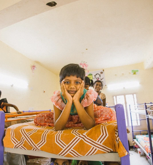 A girl sitting on her bed, hands on her face, looking at the camera with a smile