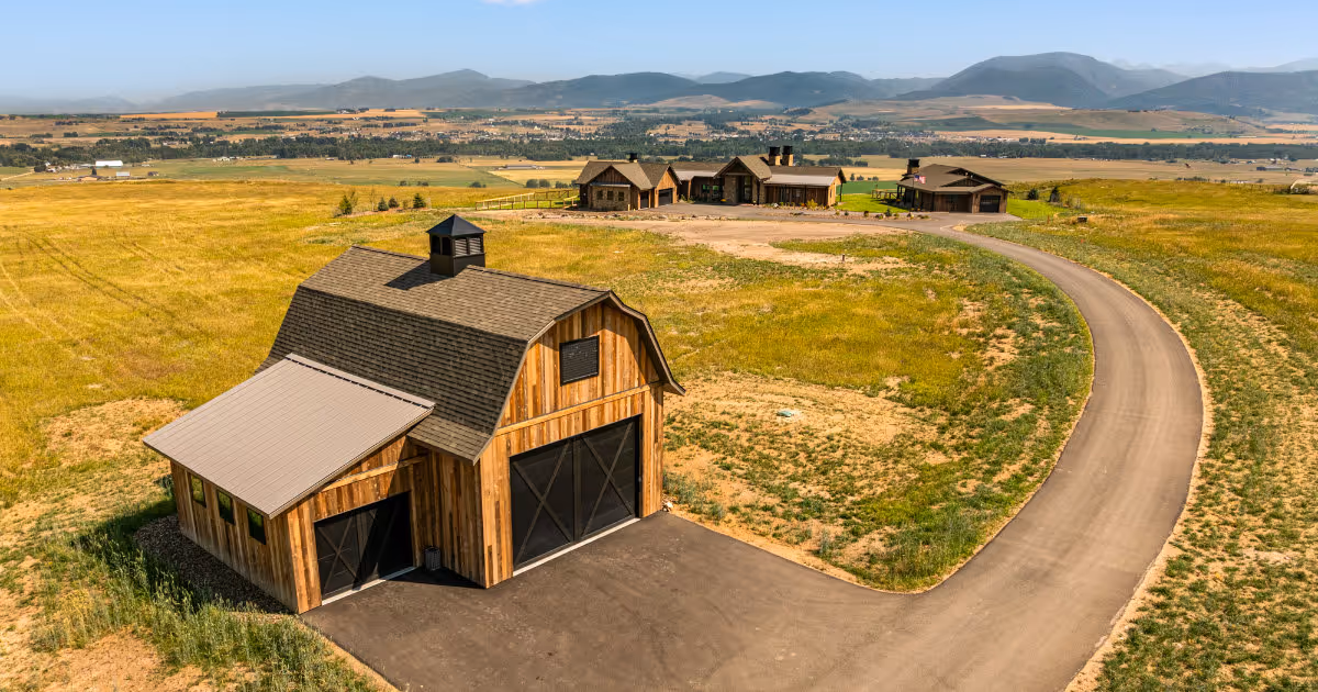 Curved paved driveway leading to a large wooden barn with a shingled roof, with three rustic houses in the background and mountains in the distance.