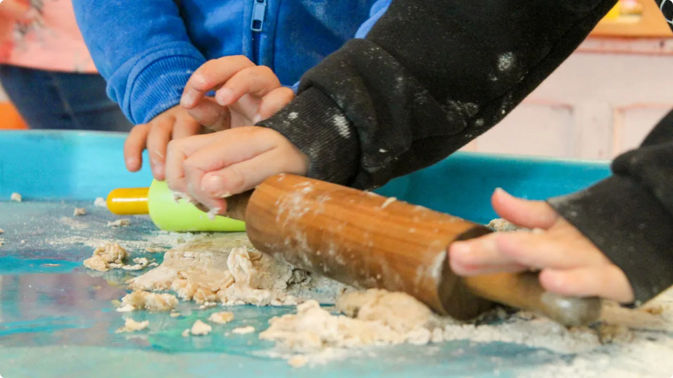 Two children rolling out dough with a wooden rolling pin on a blue surface.
