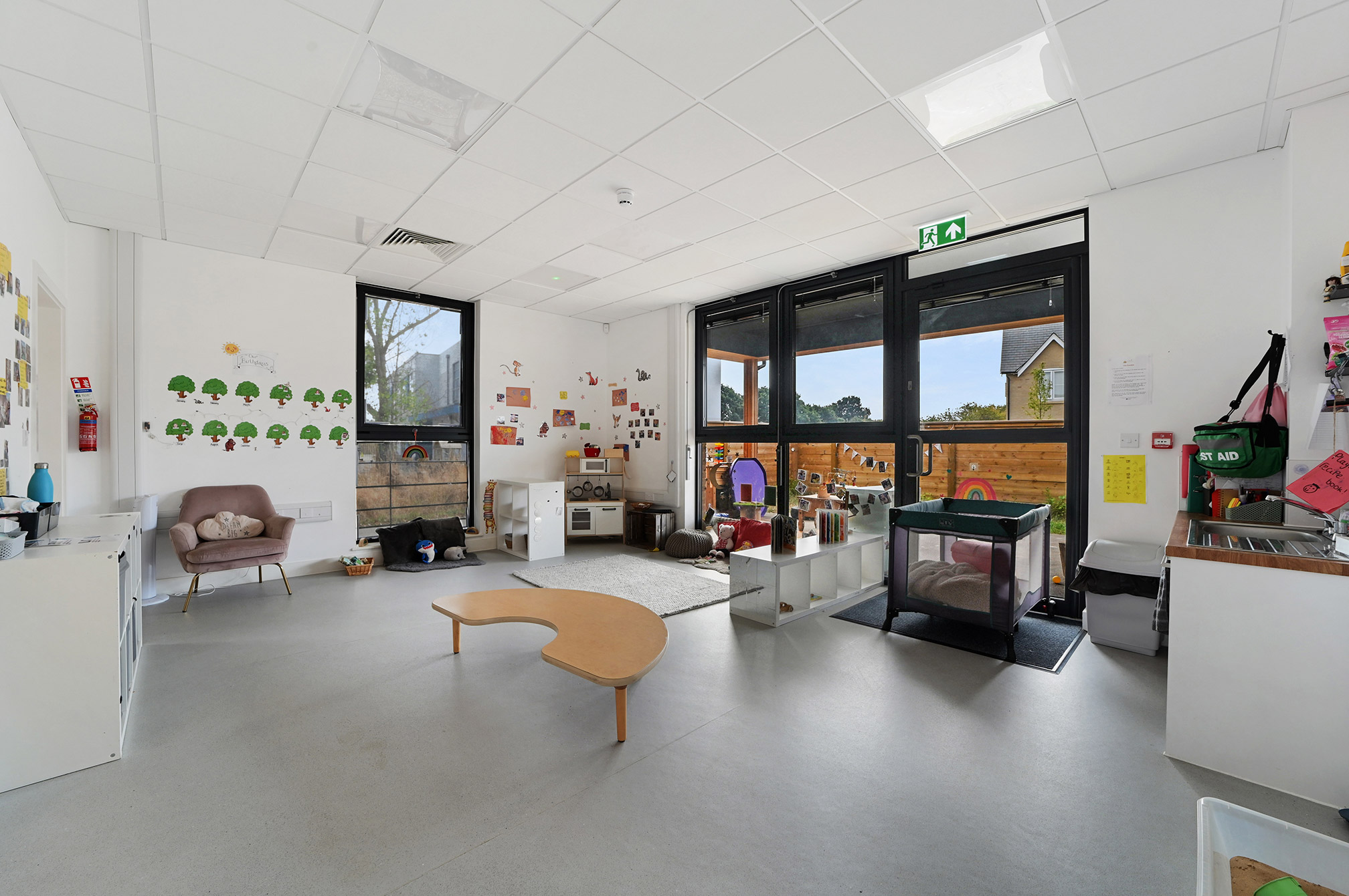 Bright and spacious children's playroom with a low wooden table, soft chair, toys, a playpen, colorful wall decorations, and large windows overlooking an outdoor play area.