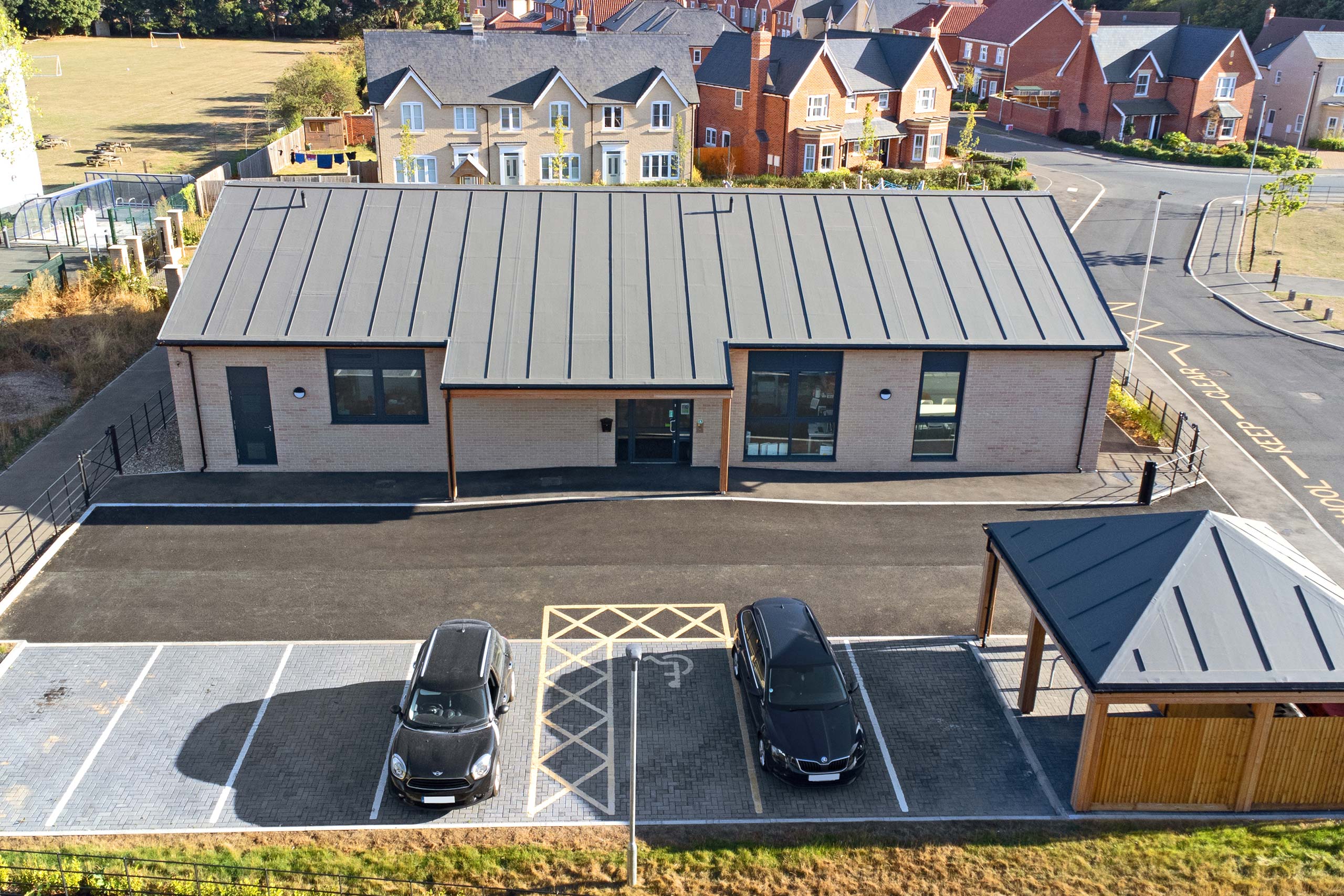 Aerial view of a modern single-story building with a gray metal roof, two black cars parked in front near a handicap parking space, and neighboring houses in the background.