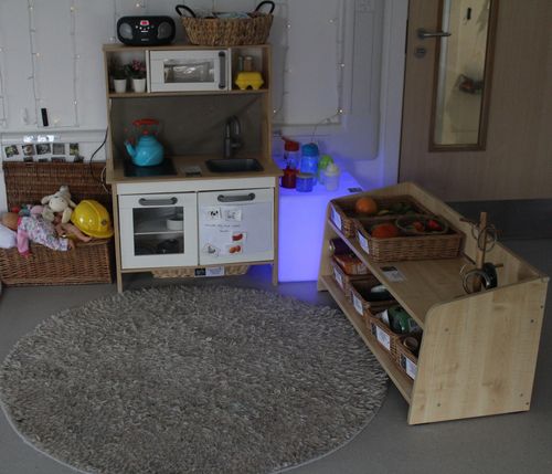 Children's play kitchen area with toy stove, sink, blue kettle, wicker baskets with play fruits, dolls, and a gray round rug.