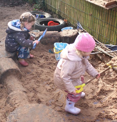 Two children wearing winter clothes playing with shovels and buckets in a sandy outdoor sandbox.