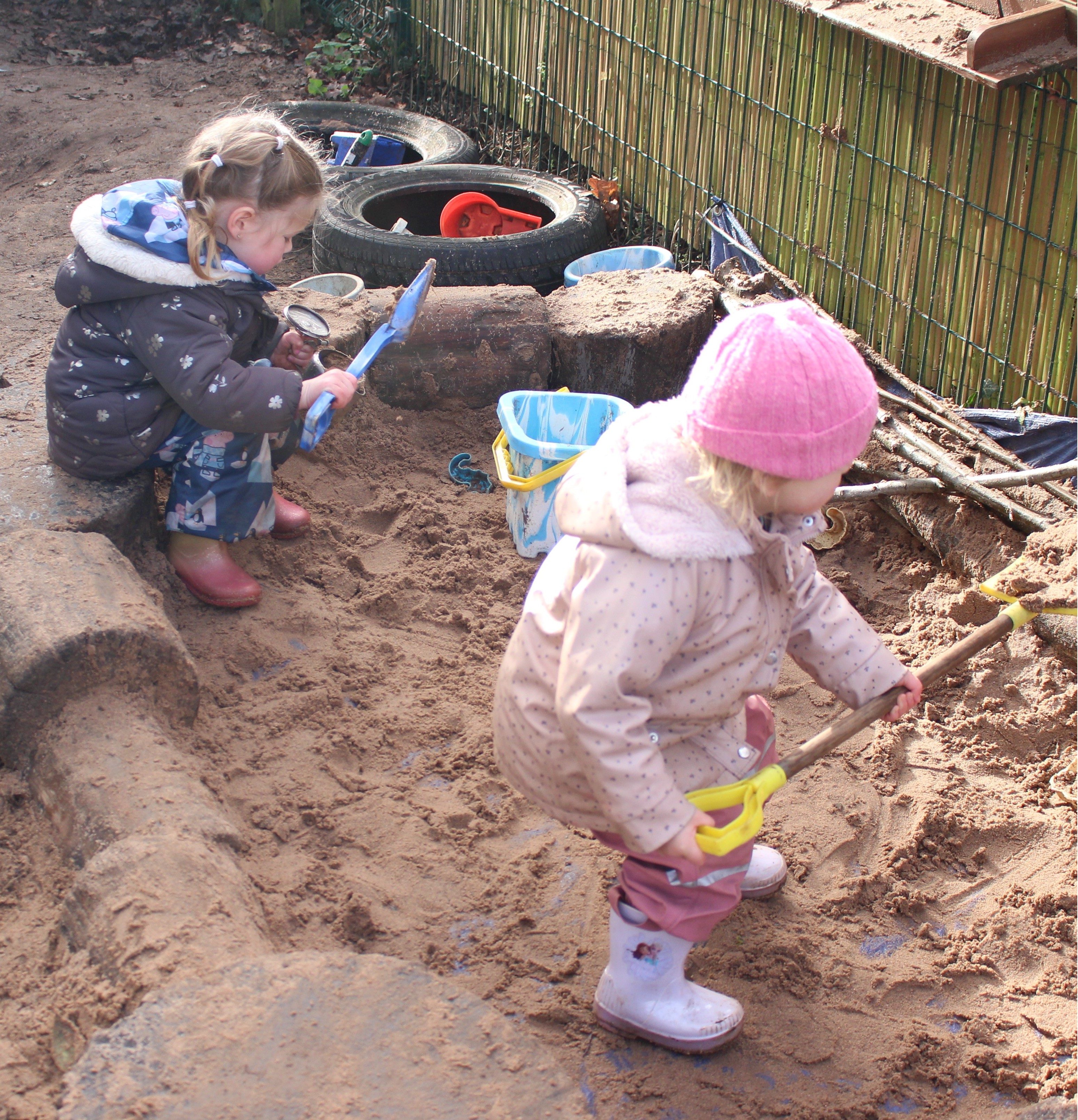 Two children wearing winter clothes playing with shovels and buckets in a sandy outdoor sandbox.