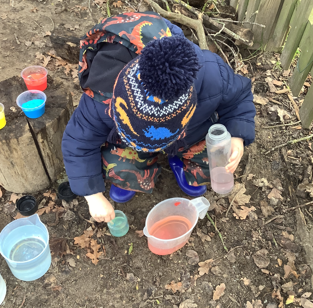 Child in dinosaur-themed clothing and knitted hat mixing colored water in plastic containers outdoors on dirt ground.