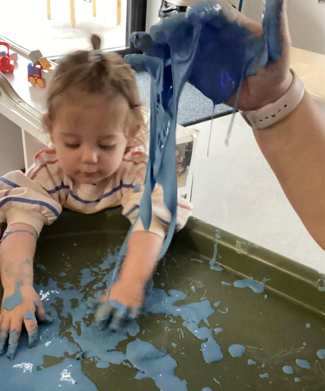 Toddler playing with blue slime on a tray, with an adult's hand holding and stretching the slime above.