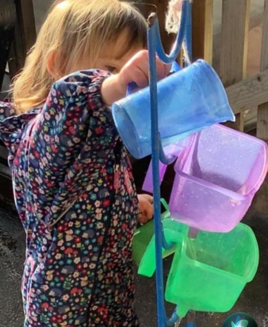 Child wearing a floral jacket pouring water from a blue cup into a series of hanging plastic containers outdoors.