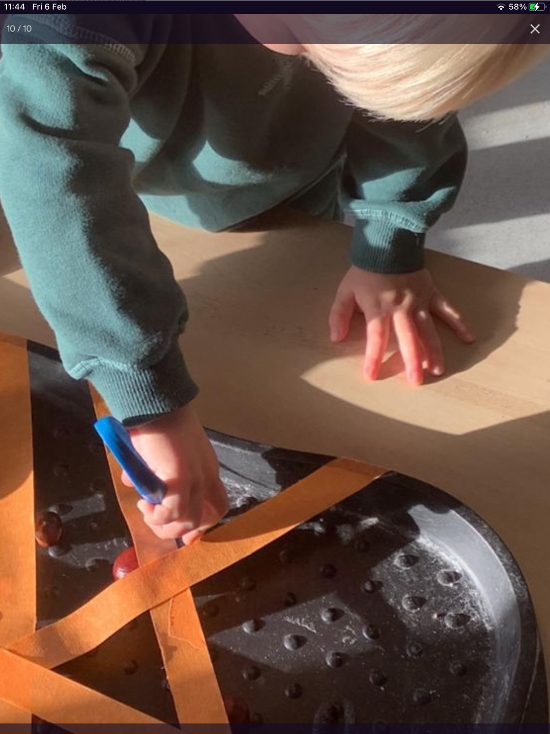 Child with blonde hair wearing a green sweater using a blue tool to poke grapes on a black tray with orange tape straps.