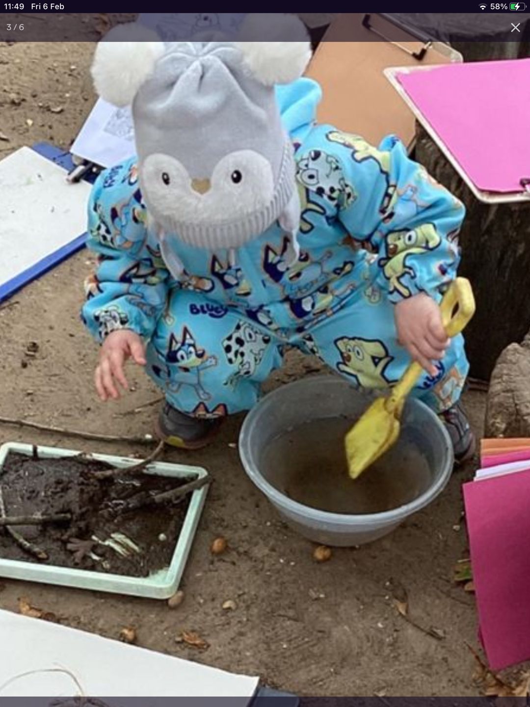 Child wearing a blue cartoon animal outfit and owl hat stirring water in a gray bowl with a yellow plastic shovel outdoors.
