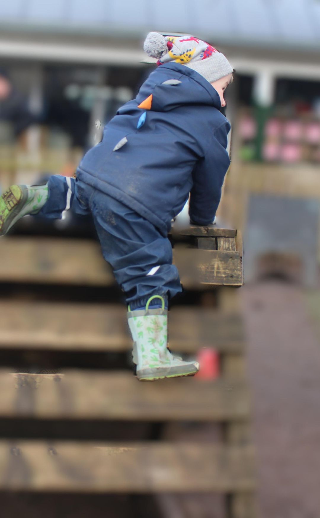 Child dressed in a blue jacket and dinosaur hat climbing on wooden pallets wearing green rain boots.