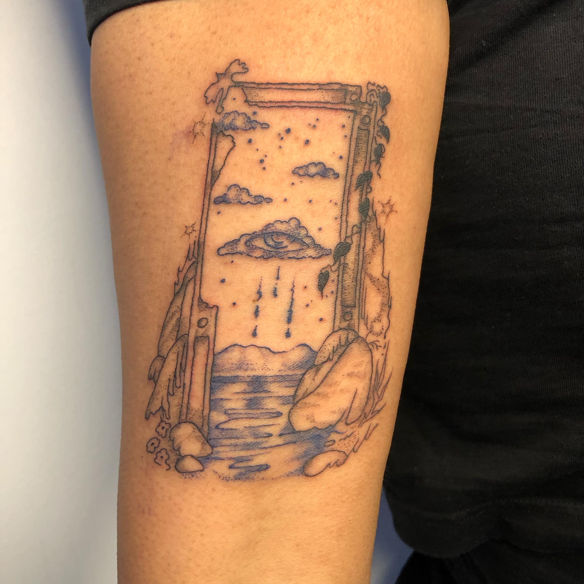 ‎a tattoo of a makeshift rotting door that is a portal to a beach with a cloud above it with a single eye and raindrop tears the door is surrounded by rocks stars and foliage its done in cobalt and black ink and the wearer of the tattoo has a medium dark skin tone