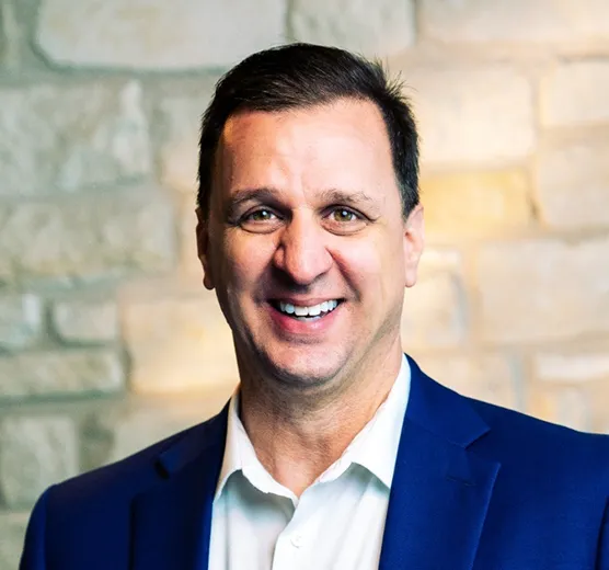 Man smiling wearing a blue blazer and white shirt against a stone wall background.