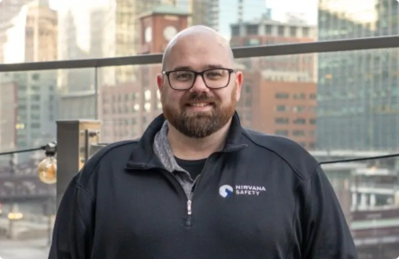Smiling bald man with glasses and a beard wearing a black Nirvana Safety jacket standing outdoors with city buildings in the background.