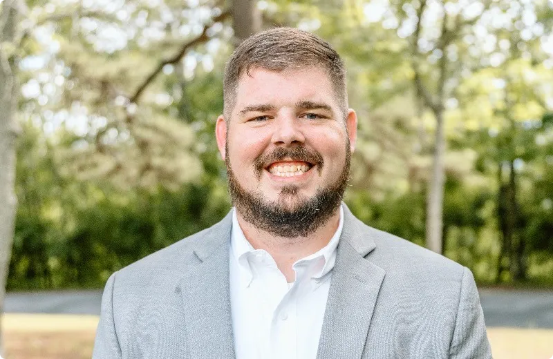 Smiling man with a beard wearing a light grey blazer and white shirt standing outdoors with trees in the background.