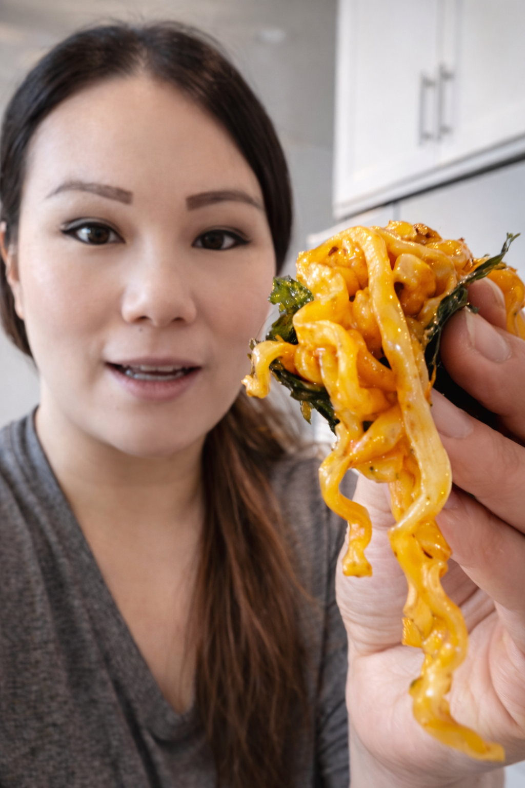 Woman holding cheesy Buldak Carbonara ramen wrapped in roasted seaweed, showing glossy spicy noodles close to camera.