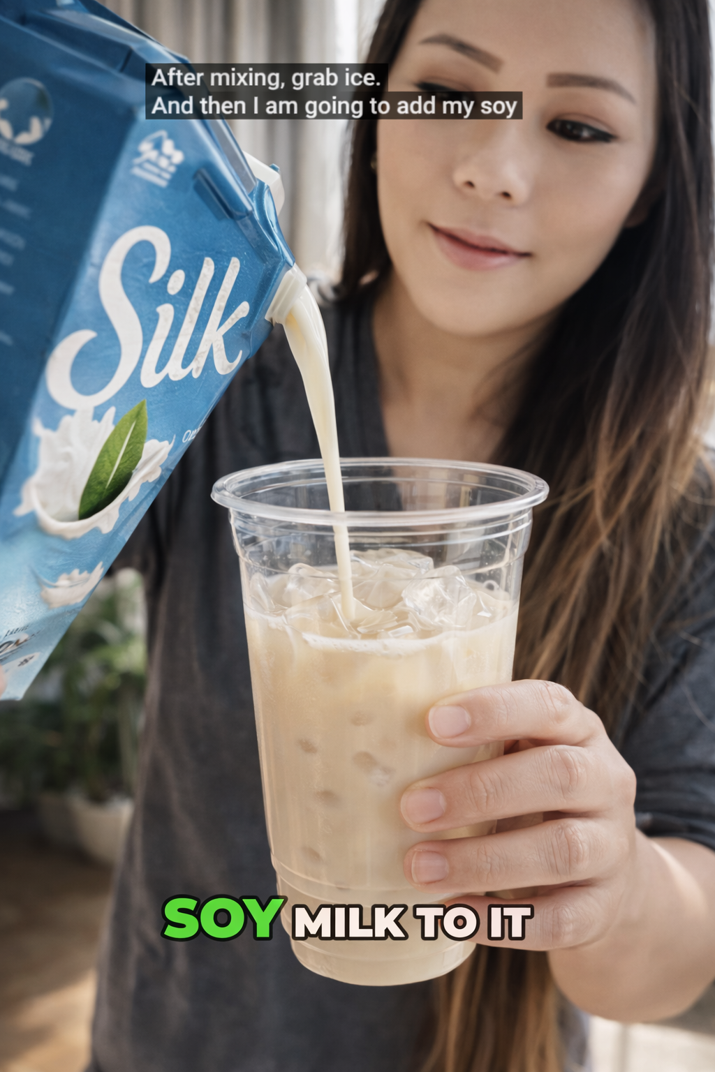 Woman pouring soy milk from a blue carton into a clear plastic cup filled with iced coffee, with visible milk stream and ice cubes, softly lit by natural window light in a cozy kitchen.