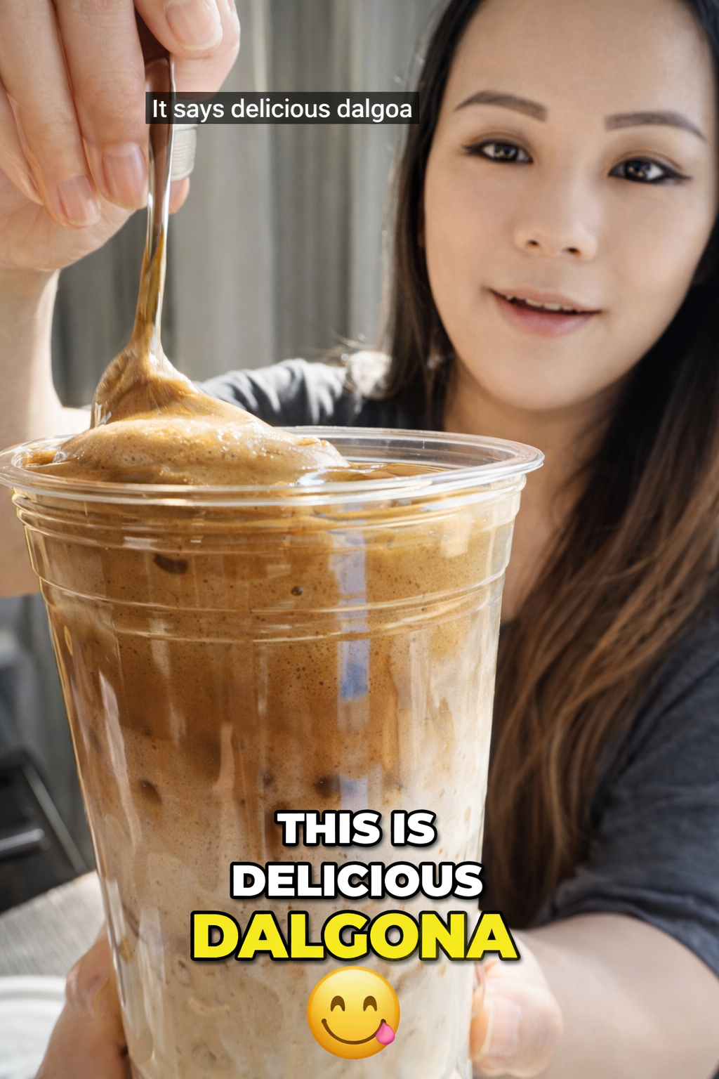 Woman holding a clear plastic cup of iced dalgona coffee topped with thick whipped coffee foam, lifting a spoonful of creamy foam, with visible milk and coffee layers in soft natural window light.