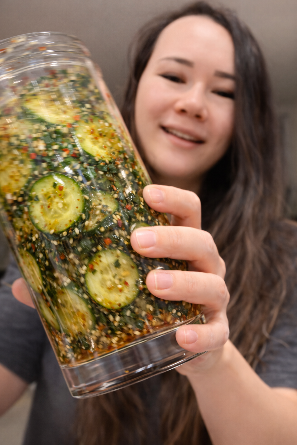 Close-up of a glass jar filled with marinated cucumber slices being shaken, the cucumbers coated in glossy soy-sesame dressing with sesame seeds, chili flakes, and seaweed seasoning, set against a softly blurred kitchen background.