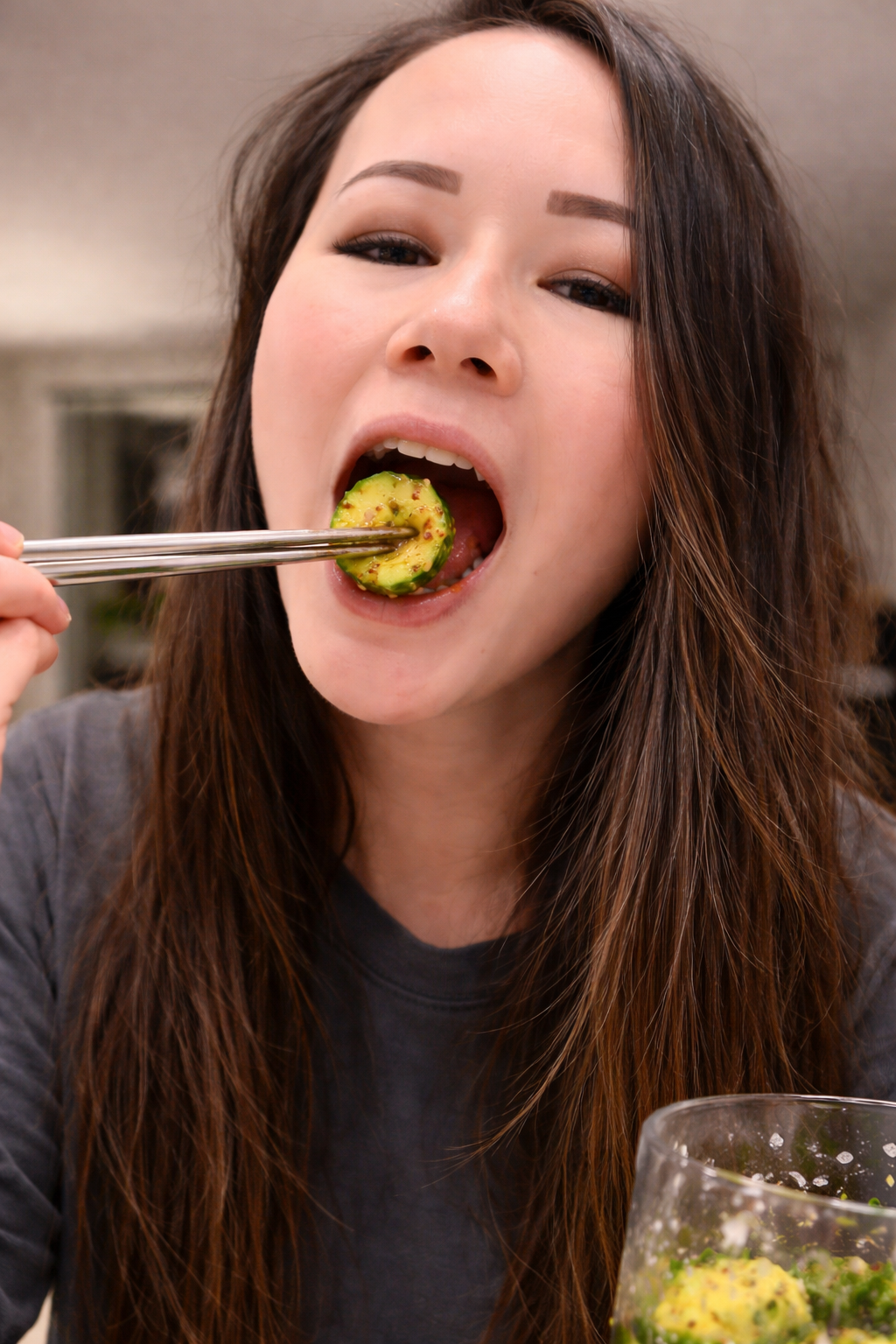 Close-up of a woman eating a marinated cucumber slice with chopsticks, the glossy cucumber coated in sesame seeds and chili flakes in sharp focus against a softly blurred kitchen background.