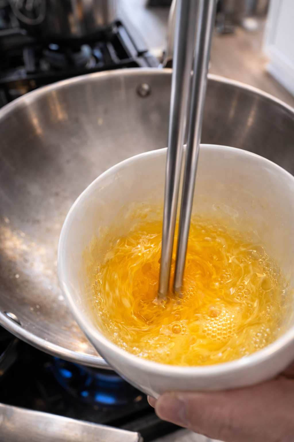 Close-up of a bright yellow egg being whisked with stainless steel chopsticks in a white bowl, with soft bubbles forming on the surface and a blurred stovetop and wok in the background.