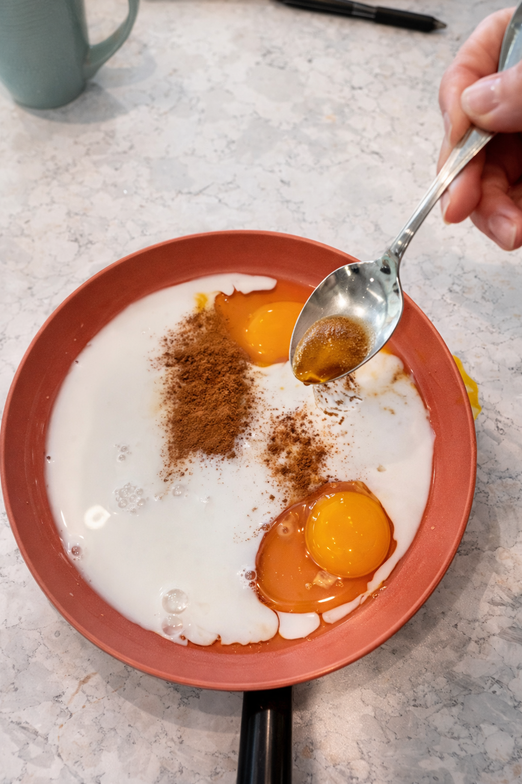 Overhead view of eggs, milk, honey, and cinnamon in a red pan on a marble countertop, with a hand holding a spoonful of honey above the mixture.