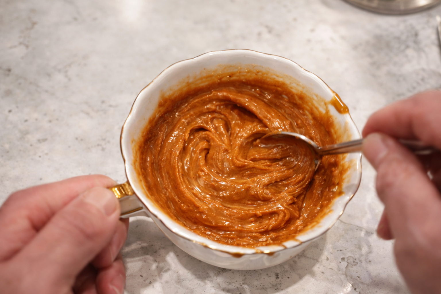 Close-up of creamy peanut butter sauce being stirred in a white bowl with a gold handle, the smooth, glossy mixture swirling richly on a marble countertop.