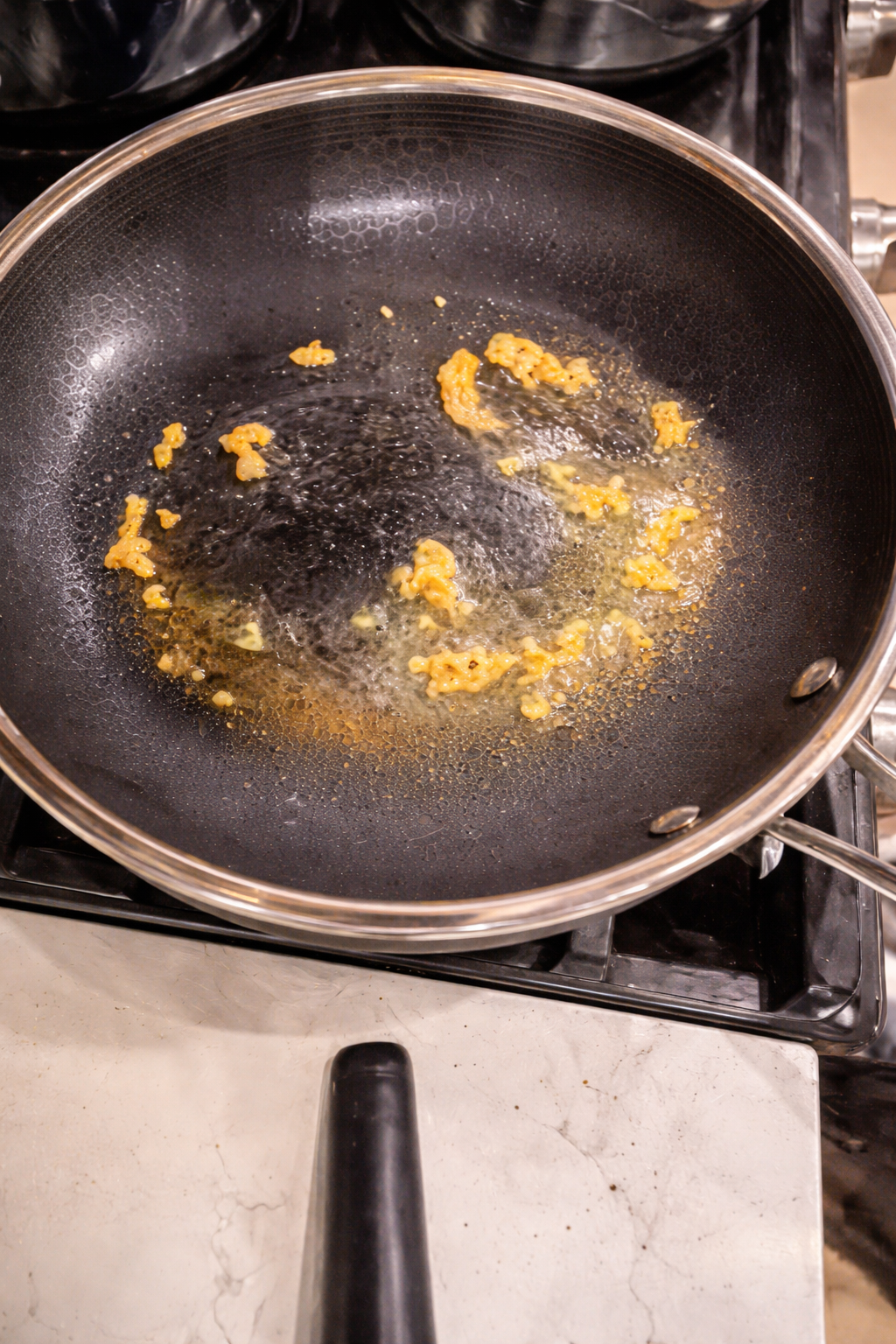 Close-up of minced garlic sizzling in olive oil inside a nonstick skillet on a stovetop, the golden pieces gently browning and glistening under warm kitchen lighting.