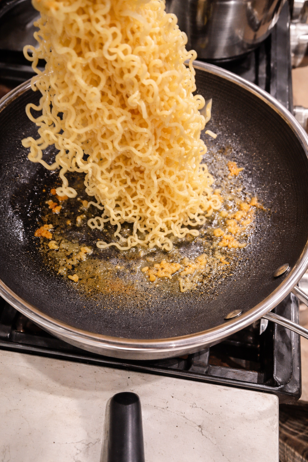 Close-up of softened ramen noodles being added to a nonstick skillet with sizzling minced garlic and olive oil on a stovetop, the noodles cascading into the pan under warm kitchen lighting.