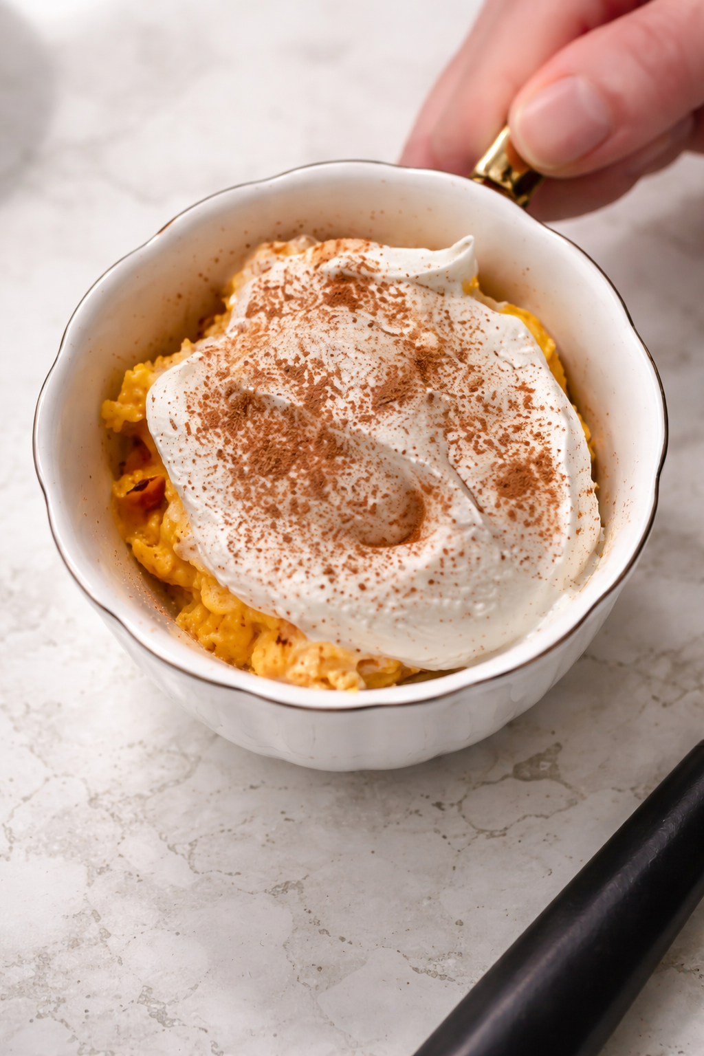 Close-up of a creamy no-bake pumpkin cheesecake in a white scalloped bowl with a gold handle, topped with a thick swirl of Cool Whip and a generous dusting of cinnamon, set on a light marble countertop.
