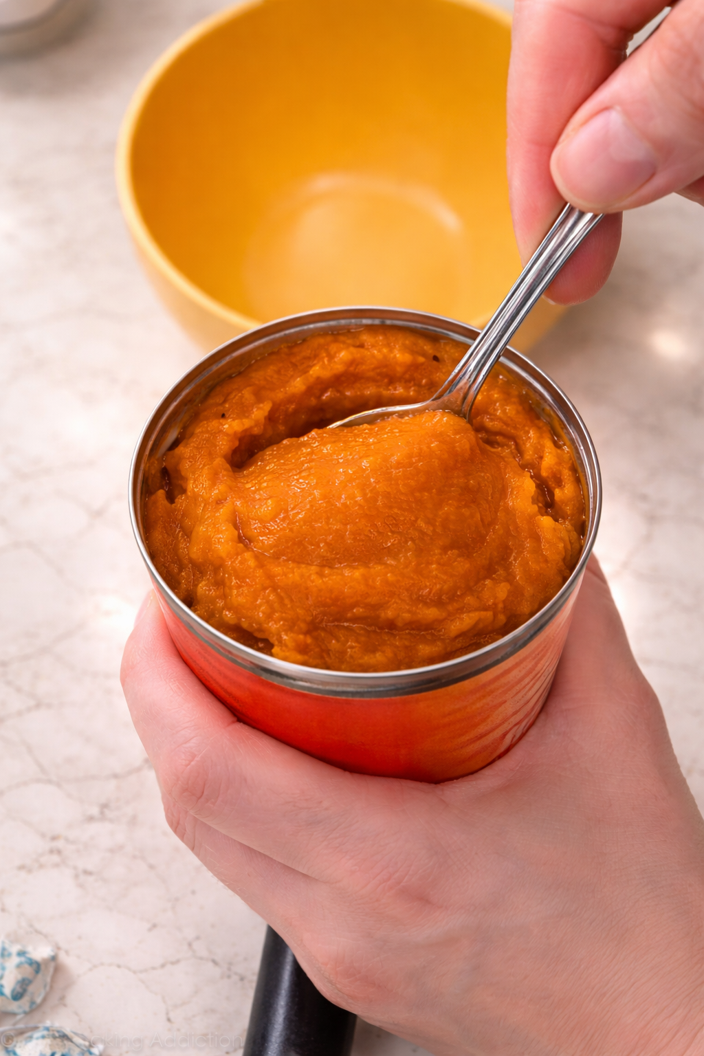 Close-up of hands holding an open can of smooth pumpkin puree while a spoon scoops the thick, vibrant orange mixture, with a yellow bowl blurred in the background on a light marble countertop.