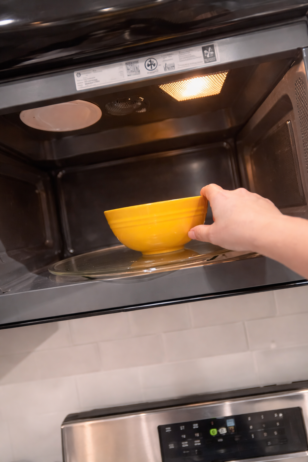 Close-up of a hand placing a bright yellow bowl into a clean stainless steel microwave, illuminated by the warm interior light, with a glass turntable visible inside.