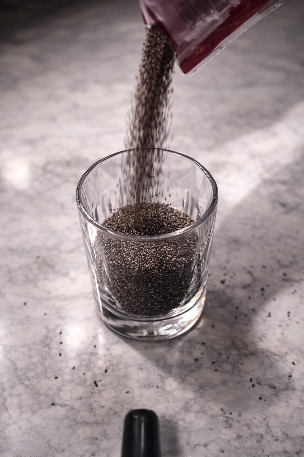 Chia seeds pouring from a package into a clear glass on a marble countertop with soft professional kitchen lighting.
