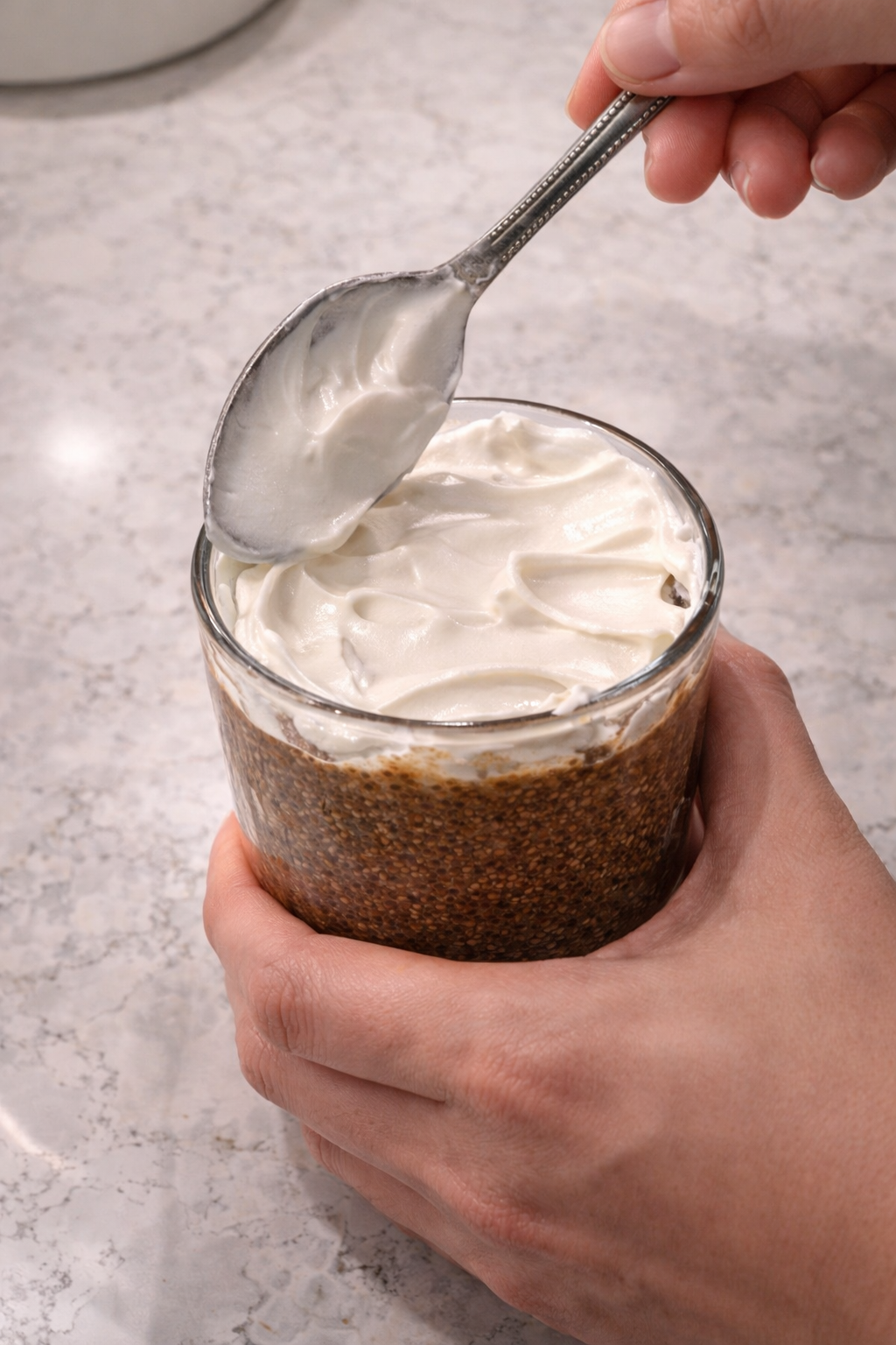 Hand spreading a thick layer of Cool Whip over a glass of tiramisu chia pudding on a marble countertop under bright professional kitchen lighting.