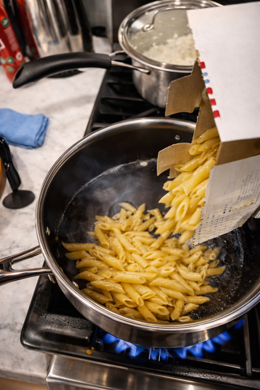 Dry penne pasta being poured from a box into a stainless steel pan on a gas stove, beginning the pasta cooking process in a home kitchen.