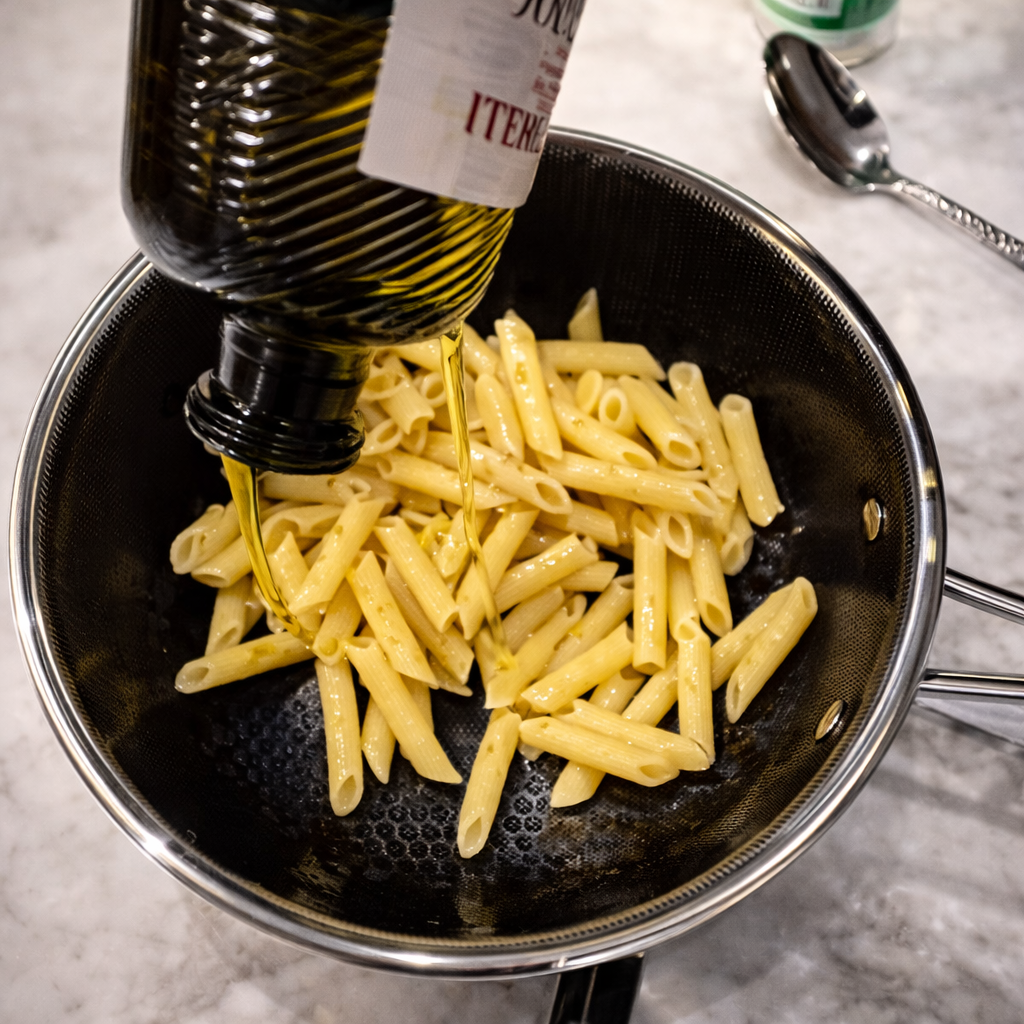 Olive oil being poured over cooked penne pasta in a stainless steel pan, preparing the pasta for seasoning while it glistens under bright kitchen lighting.