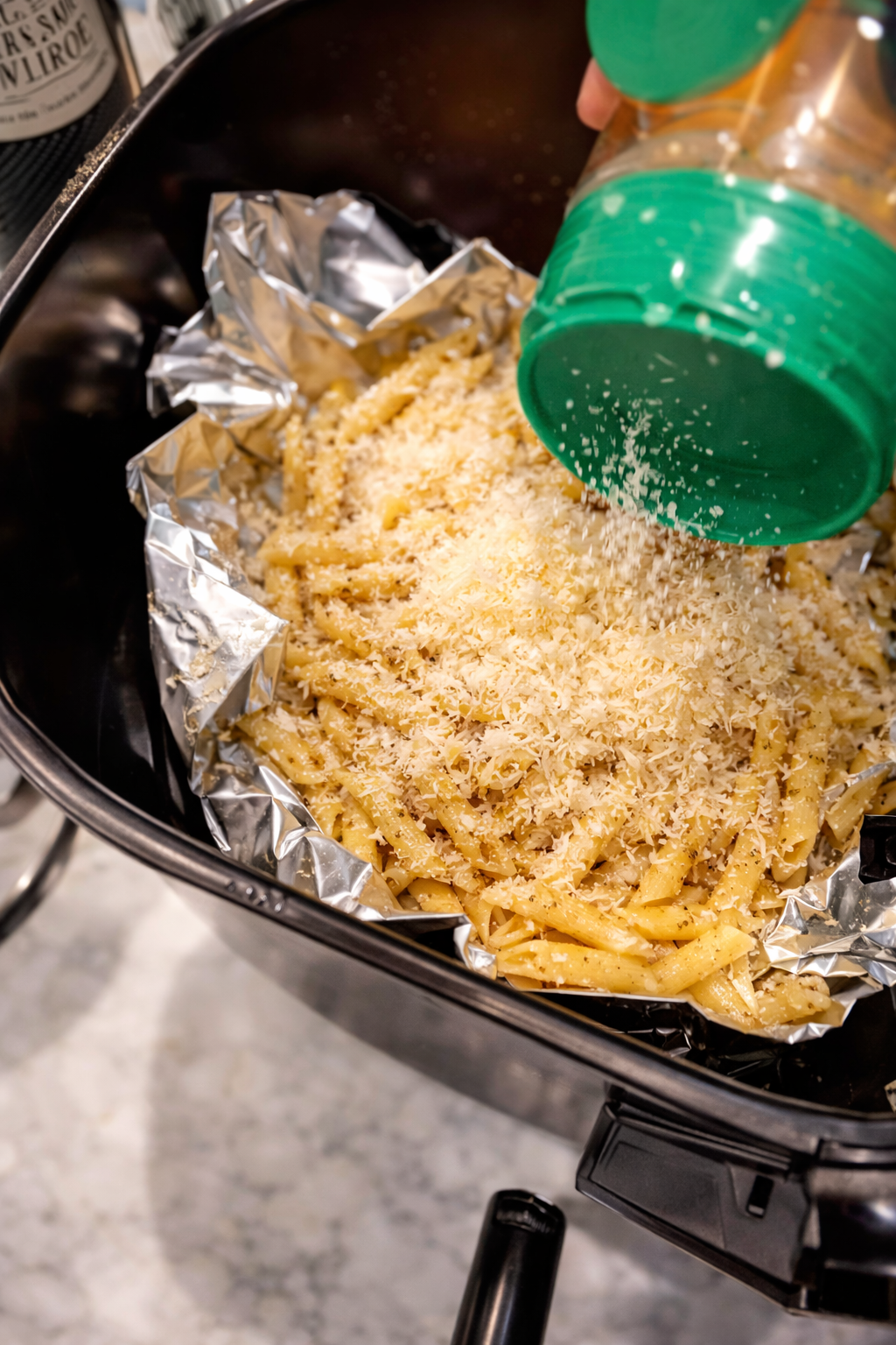 Grated Parmesan cheese being sprinkled over seasoned penne pasta in a foil-lined air fryer basket, coating the pasta before baking into crispy pasta chips.