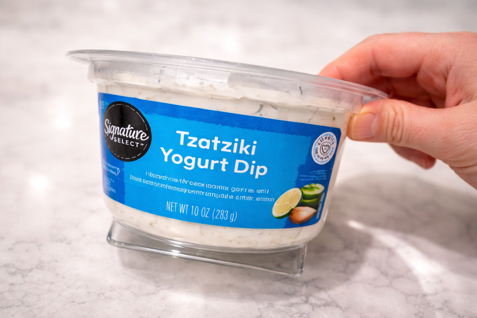 Hand holding a container of tzatziki yogurt dip on a marble countertop, creamy cucumber dip used as a sauce for crispy pasta chips.