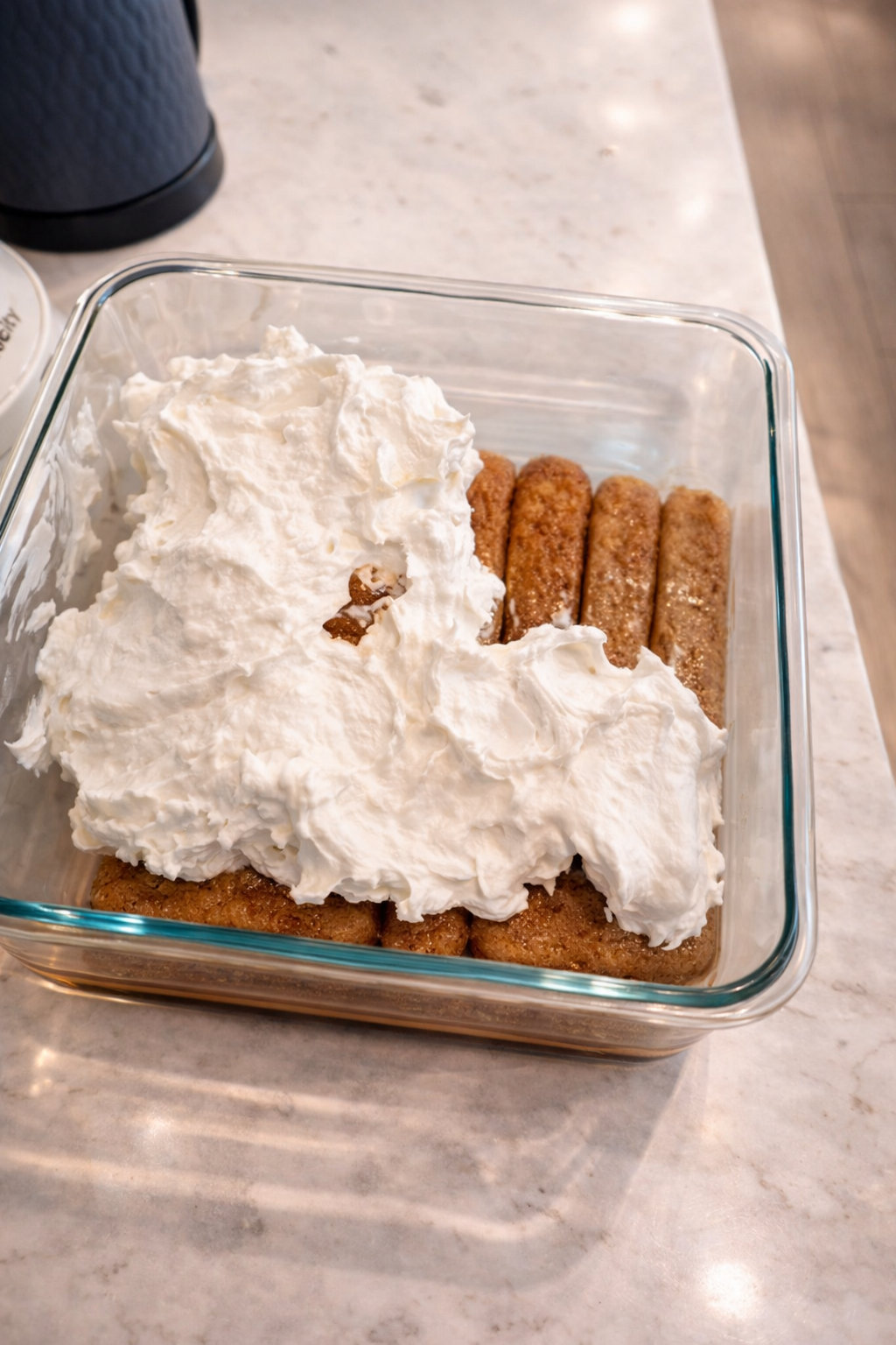 Creamy whipped topping spread over coffee-soaked ladyfingers in a glass baking dish while assembling a tiramisu dessert.