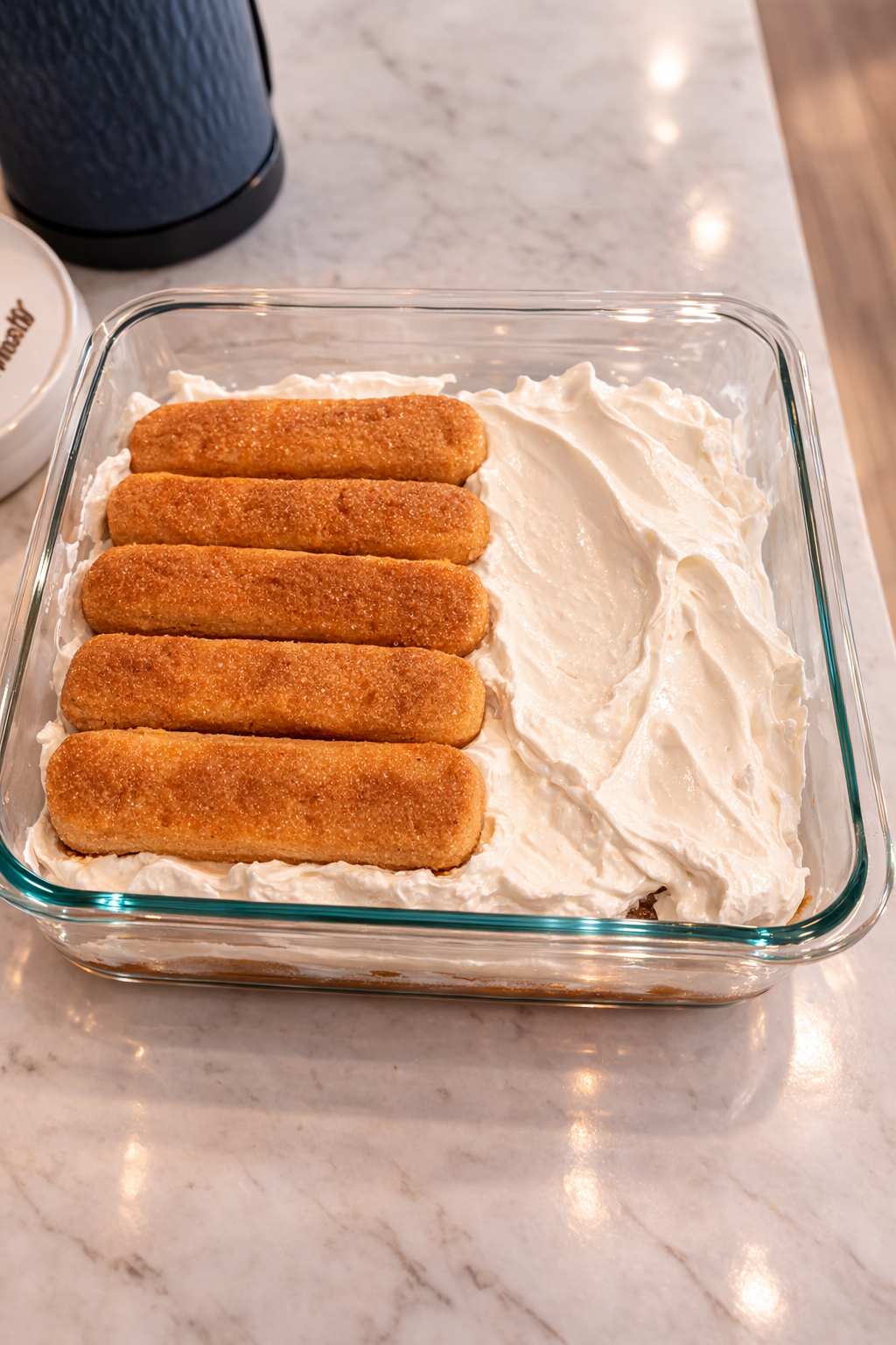 Second layer of tiramisu being assembled with ladyfinger cookies placed over a creamy whipped mixture in a glass baking dish on a kitchen countertop.