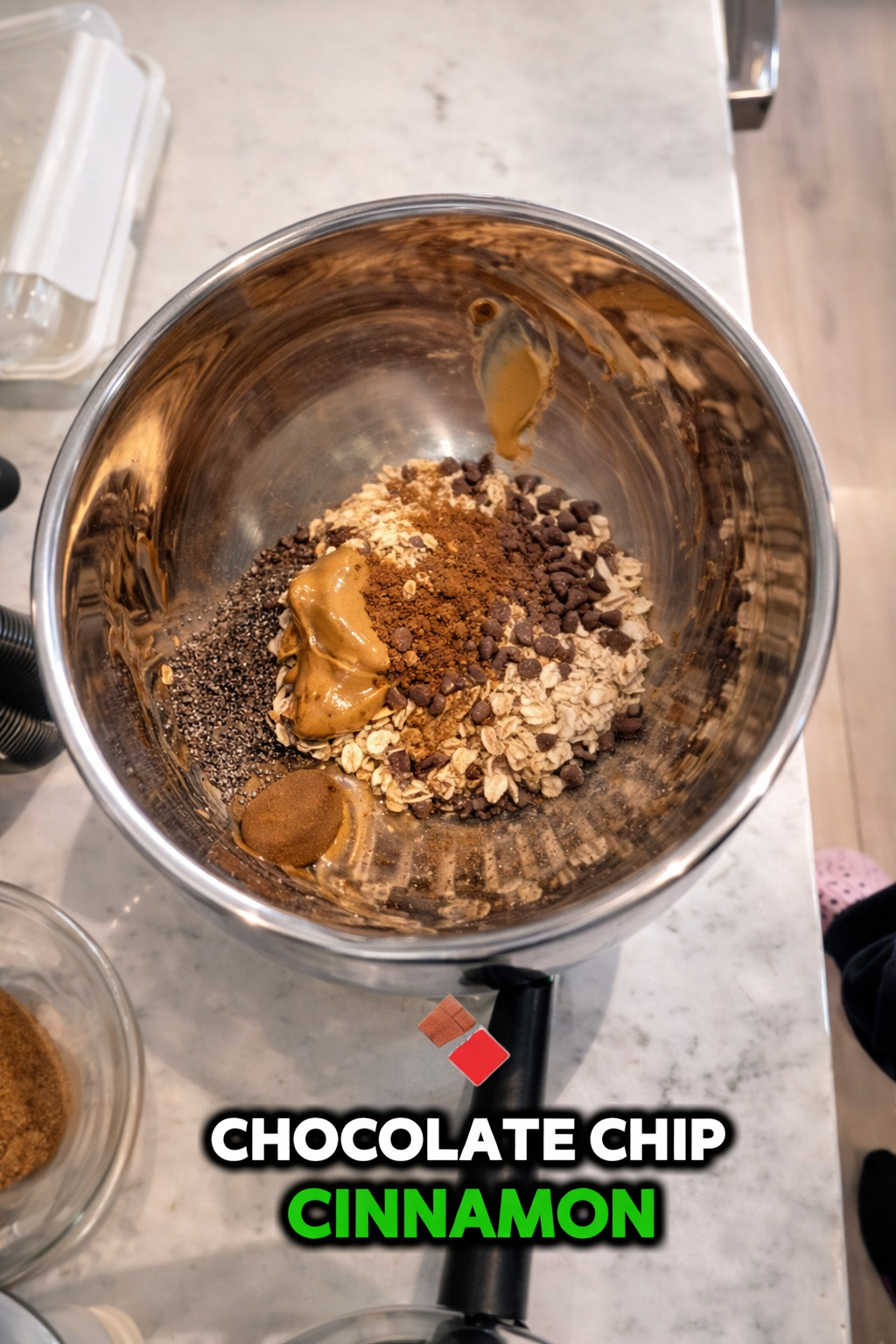 Overhead view of a stainless steel mixing bowl containing oats, chia seeds, peanut butter, chocolate chips, and cinnamon, captured with soft professional lighting that enhances the rich textures and warm tones of the ingredients on a marble countertop, with labeled text highlighting chocolate chips and cinnamon.