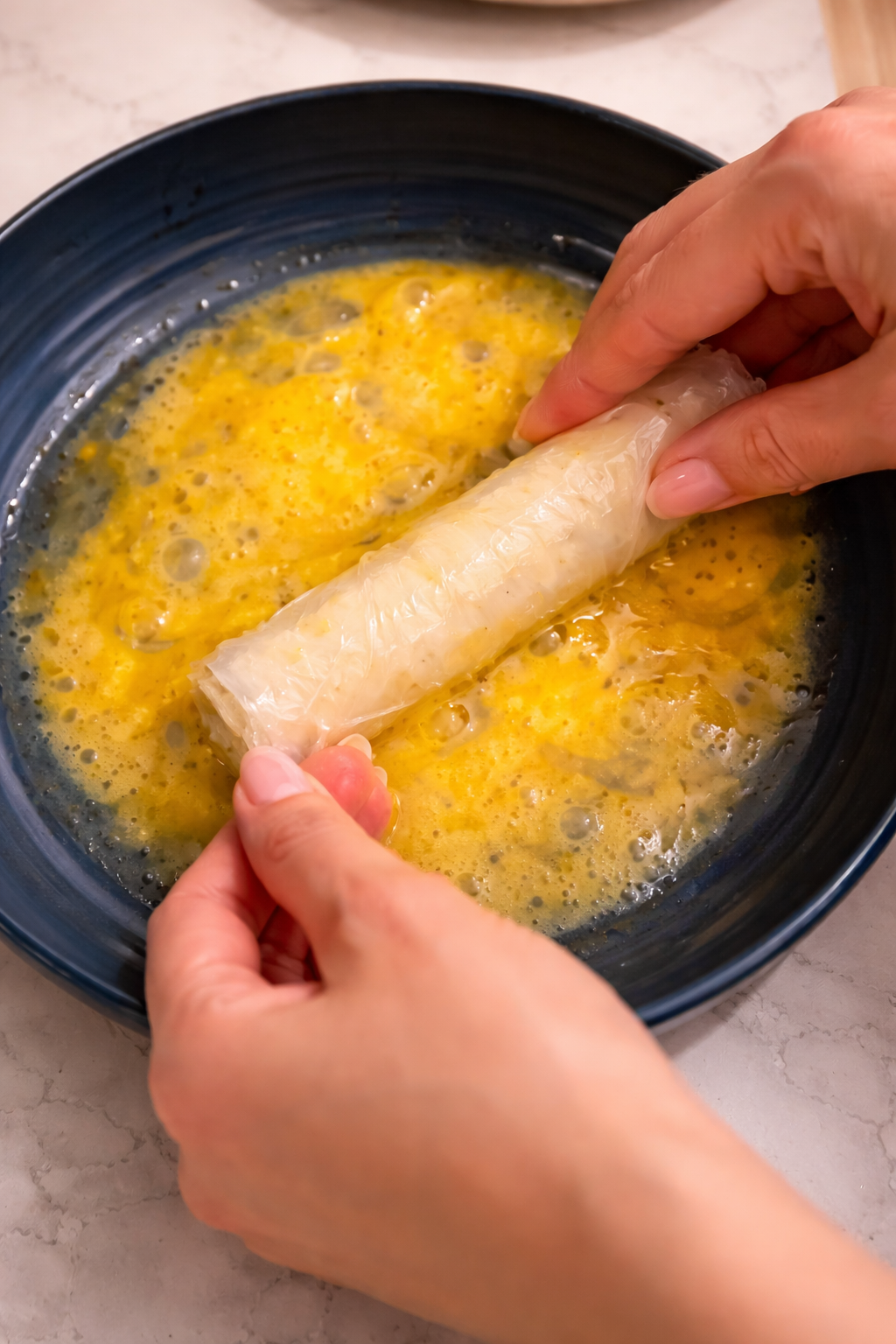 Hands dipping a rice paper-wrapped mozzarella stick into a bowl of beaten eggs on a marble countertop under bright, professional lighting.