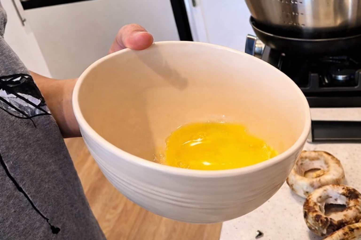 Close-up of a person holding a white ceramic bowl with melted butter inside, in a kitchen setting with a stovetop and browned mushroom caps on the counter nearby.