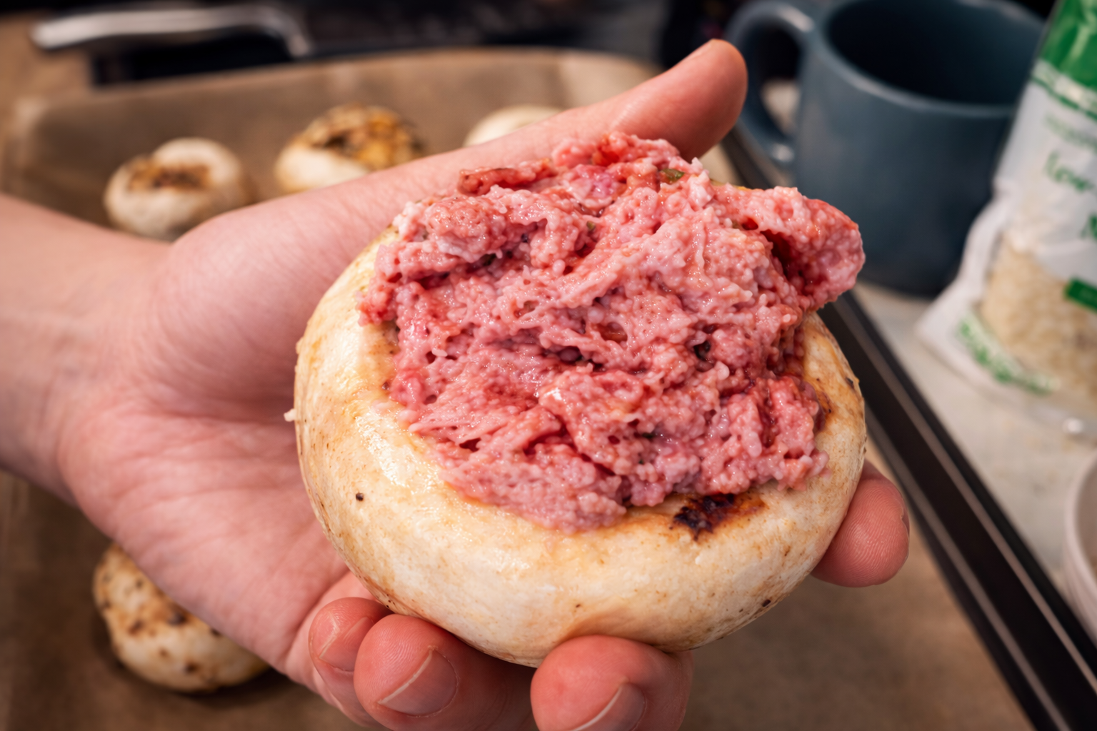 Close-up of a hand holding a large white mushroom cap filled with seasoned ground meat mixture, with additional mushrooms on a baking tray and kitchen items softly blurred in the background.