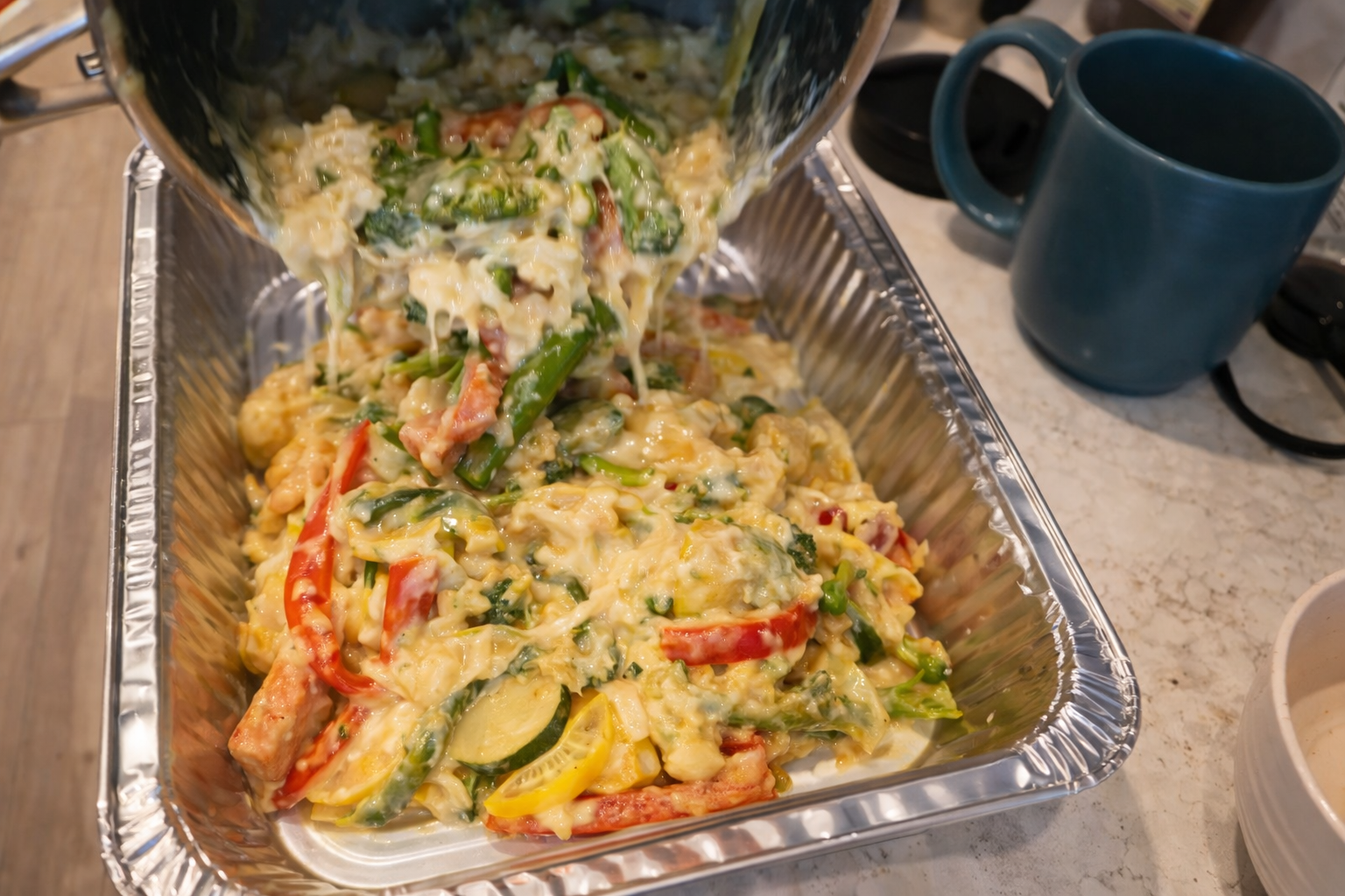 Close-up of creamy vegetable casserole being poured into a foil baking tray, with melted cheese coating broccoli, zucchini, and bell peppers under warm, professional lighting.