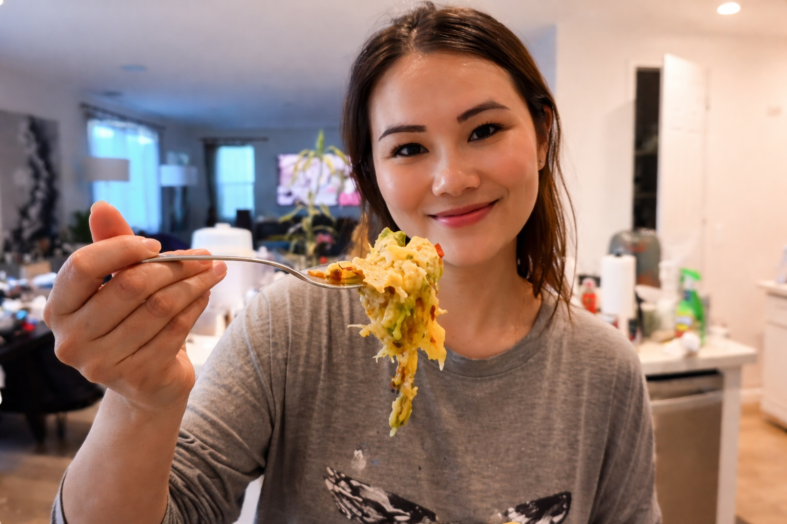 Young woman with a messy bun smiling in a modern kitchen while holding a forkful of cheesy vegetable casserole, with melted cheese stretching over mixed vegetables under bright, professional lighting.