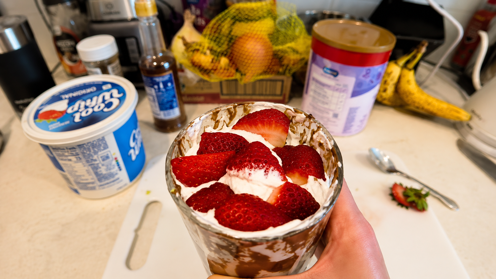 Close-up of a creamy chocolate mousse parfait topped with fresh sliced strawberries and whipped cream in a glass cup, held by a hand over a softly blurred kitchen countertop with dessert ingredients in the background.