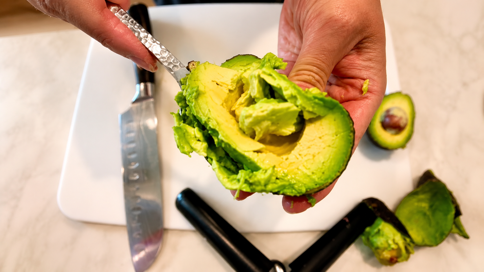Close-up overhead photo of hands scooping ripe avocado from its peel over a white cutting board with a chef’s knife nearby in a bright kitchen setting.