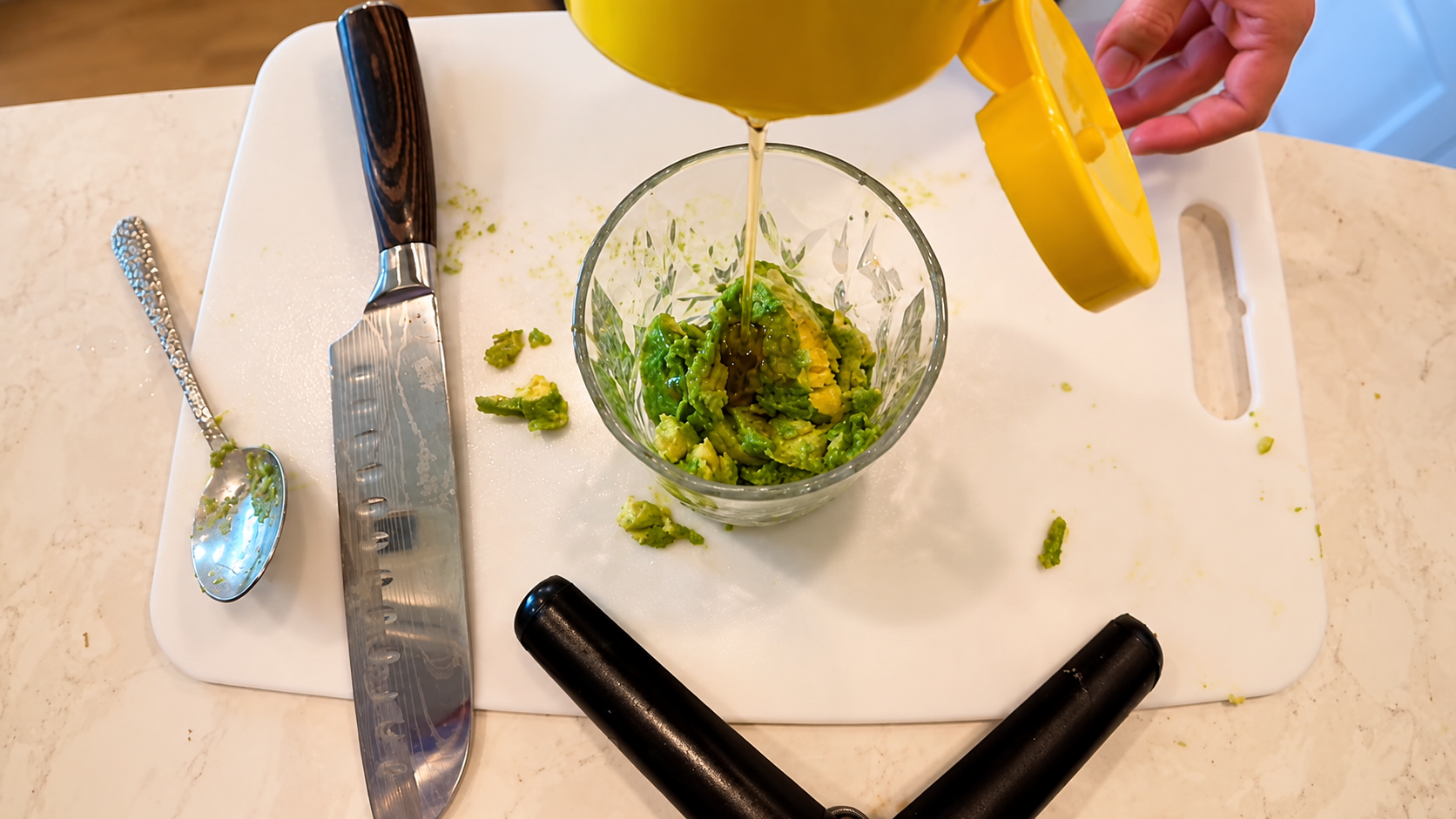 Overhead kitchen photo of honey being poured into a glass bowl filled with mashed avocado on a white cutting board beside a chef’s knife and spoon.